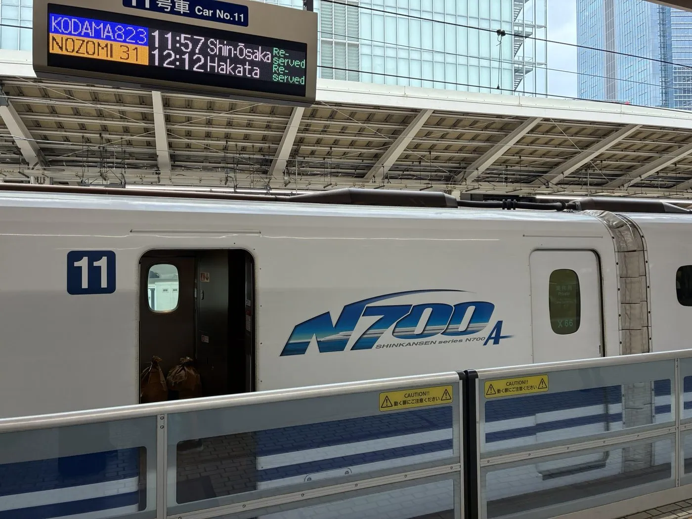 A Shinkansen N700A train at a station platform, under a departure board showing Kodama and Nozomi services to Shin-Osaka and Hakata